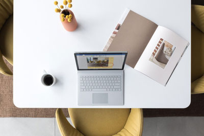  Top view of a Surface Book 2 on a tea table, with a cup of coffee, a brochure and a vase with flower in on the table  