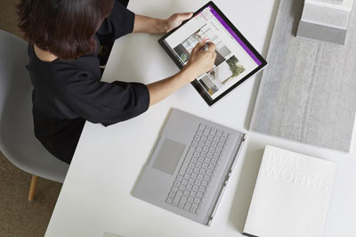  A female sitting in front of a desk drawing on a Surface Book 2 with a Surface Pen. The keyboard is removed and placed on the desk  