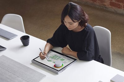  A female sitting in front of a desk drawing on a Surface Book 2 with a Surface Pen, with the keyboard attached and fold behind. A Surface Dial is placed on the screen  