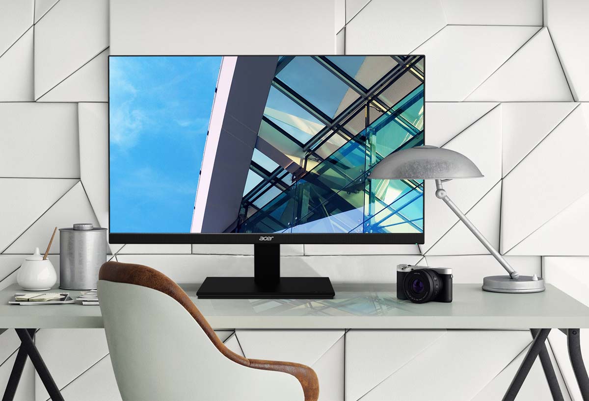 Acer V277U on a desk next to a DSLR camera, lamp, modernly stylized wall and a screenfill of the edge of windows on a modern building