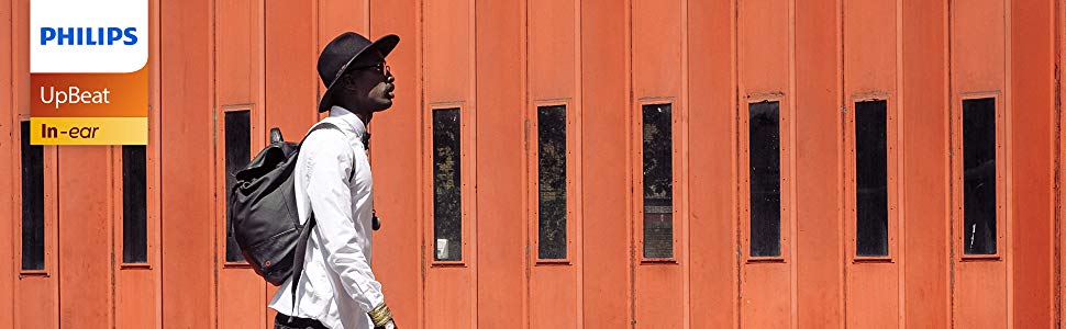 A man walking in front of a brown wall, wearing a backpack, a hat, and these earphones over his neck 