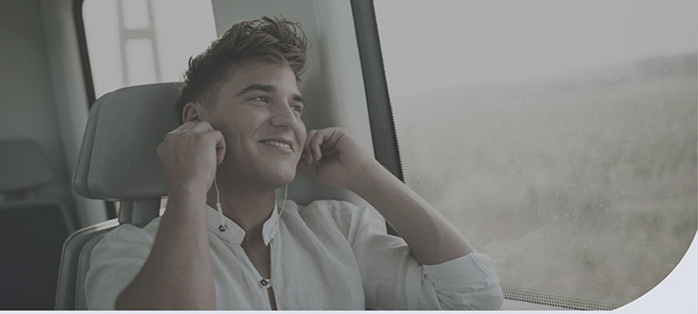 A man inside a bus, next to a window putting on his white earbuds
