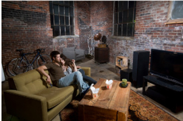 A man and woman on a couch close together, in their brick apartment angled towards their entertainment center that includes a TV and the Energy CC speakers
