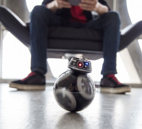 A man sitting in a chair controlling a BB-9E unit that's on the floor
