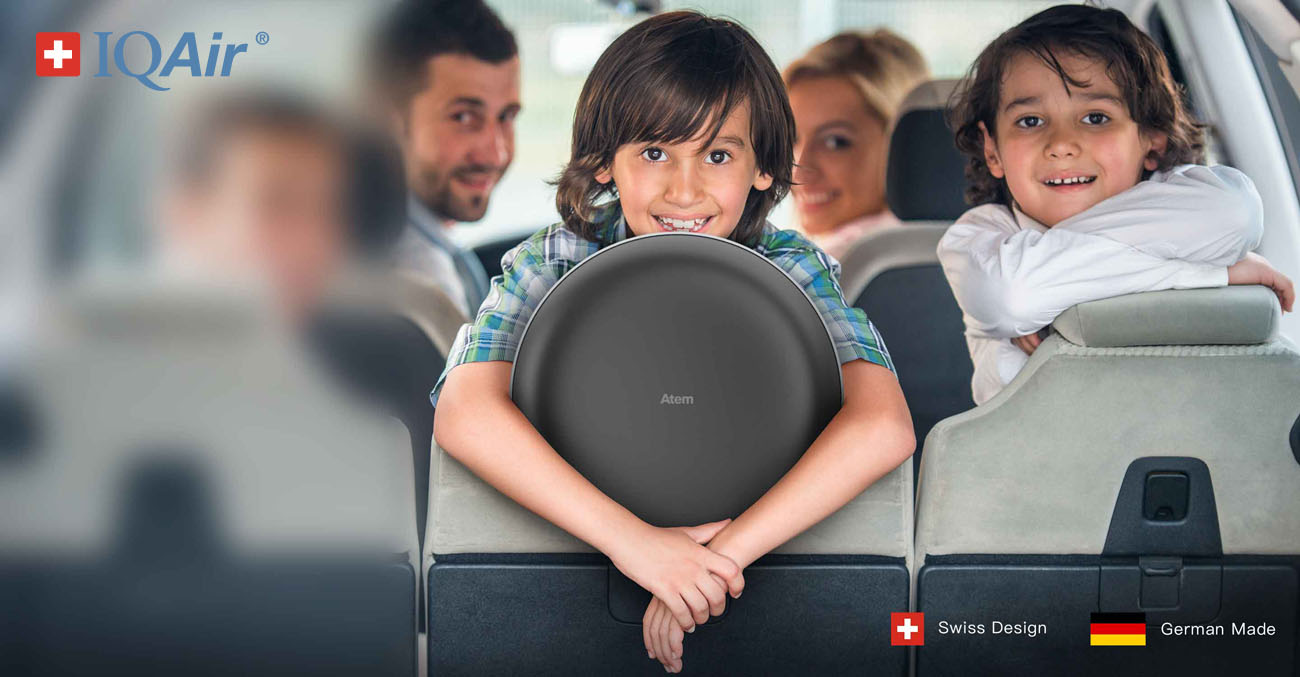 IQAir Atem Car banner showing a family in their car facing to the back of their seat smiling at the camera. The IQ Air logo, Swiss and German flags are there along with text that reads Swiss Design, German Made