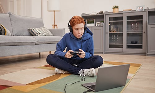 A boy is playing game on his laptop with headset over his head and controller in his hands