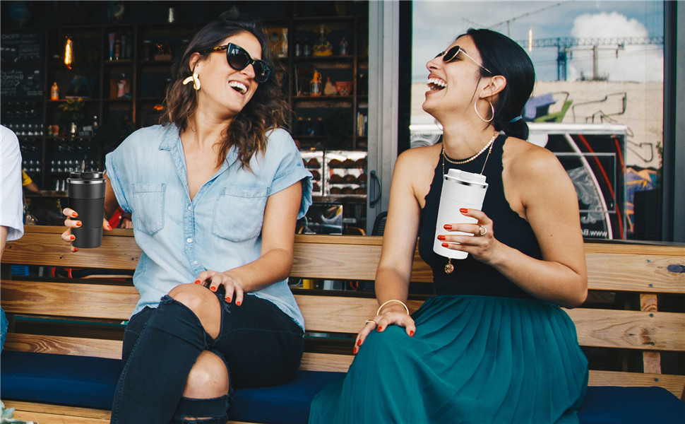 Two women sitting outdoors, laughing. One wears a blue shirt, the other holds a takeaway drink. They appear to be at a cafe.