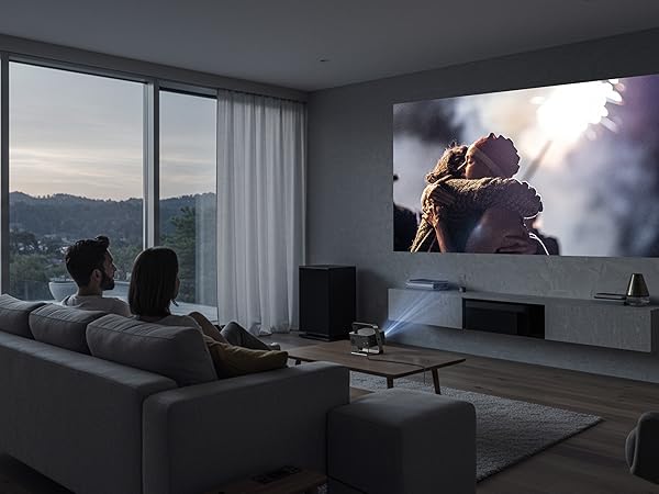 Modern living room with large windows and a giant projector screen displaying a nature scene. Two people sit on a gray couch, facing the screen.
