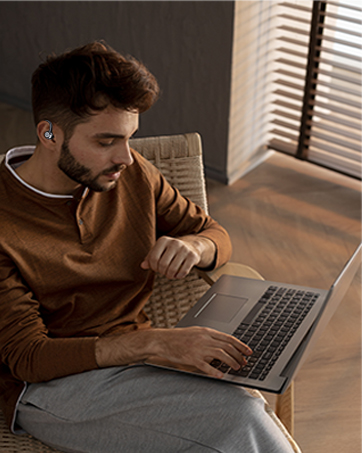 Person wearing wireless earbuds while working on a laptop. Seated in a chair near a window with blinds, wearing a brown sweater.