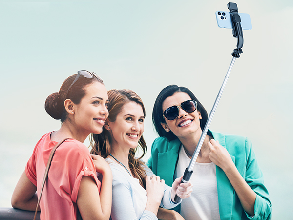 Extendable selfie stick with smartphone mount being used by three people to take a group photo outdoors.