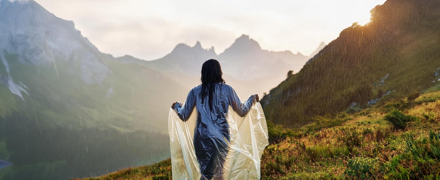 Person wearing blue jacket holds sheer fabric in mountainous landscape. Sunlit peaks, green slopes, and misty valley visible in background.