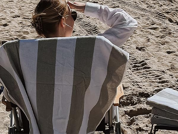 A woman sitting down at the beach with her beach towel hanging from the back of the chair.