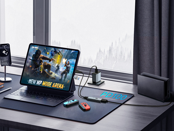 Gaming setup on desk featuring tablet with action game, colorful controllers, USB hub, and large mousepad. Window with cityscape visible in background.