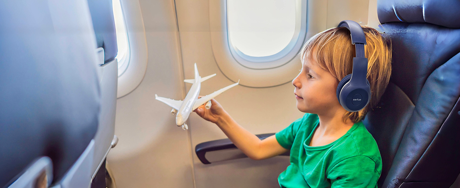 Child in green shirt on airplane seat, wearing headphones and holding small white toy airplane near window. Leather seats and aircraft interior visible.