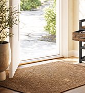 Round beige/tan woven floor mat or rug placed in home entryway near window with natural light, wooden door frame visible.
