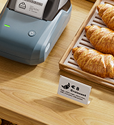 Partial view of a light blue device on wooden surface next to baked goods. Small white label visible in foreground.