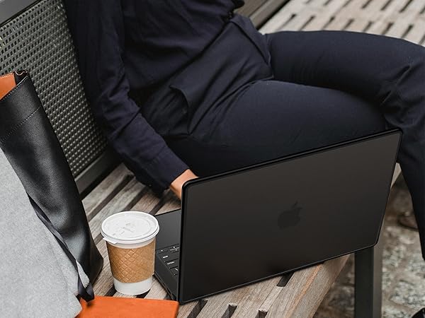 Person sitting on outdoor bench with black laptop. Coffee cup and orange notebook visible. Casual work or study scene.