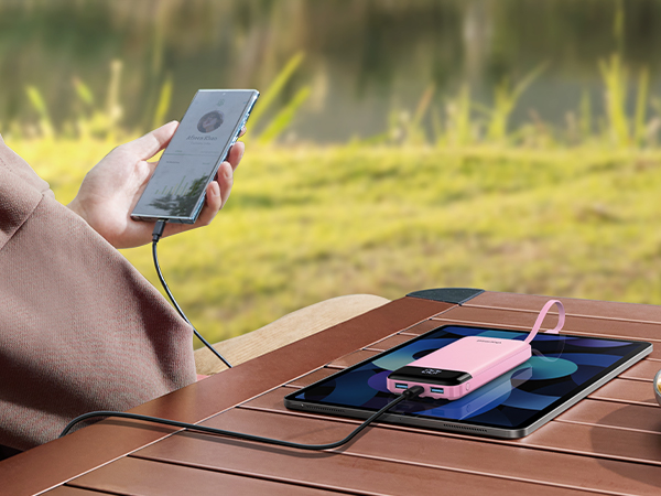 Outdoor scene with hands holding a smartphone connected to a solar charging panel on a wooden surface. Grassy background visible.