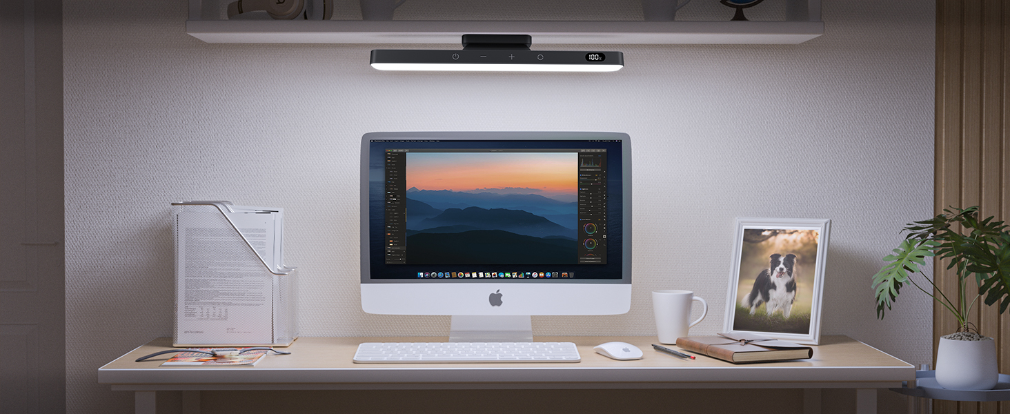 Desktop workstation with iMac computer, soundbar above screen, desk lamp, plant, and framed photo on wooden surface.