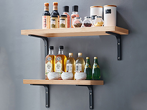 Two-tiered wooden wall shelf with black metal brackets displaying various bottles and glassware against a gray wall.