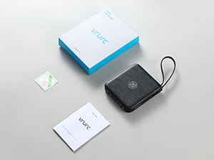 Small black electronic device with wrist strap alongside white product boxes and a small green logo sticker on a light gray surface.