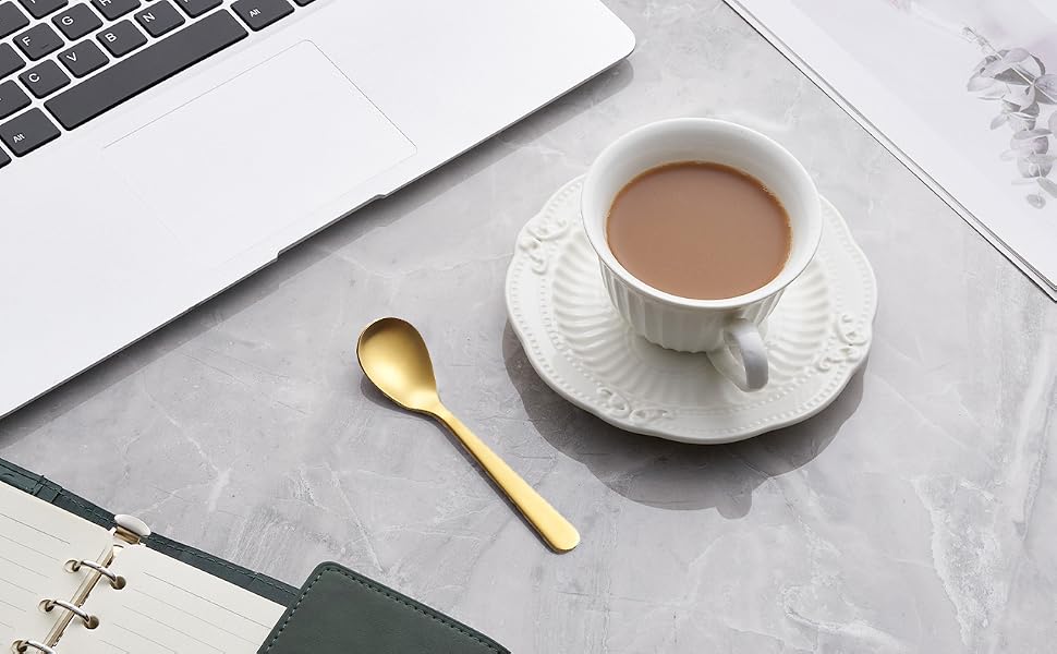 Workspace setup with laptop, white coffee cup on saucer, gold spoon, and open notebook. Clean, minimalist desktop arrangement suggesting productivity.
