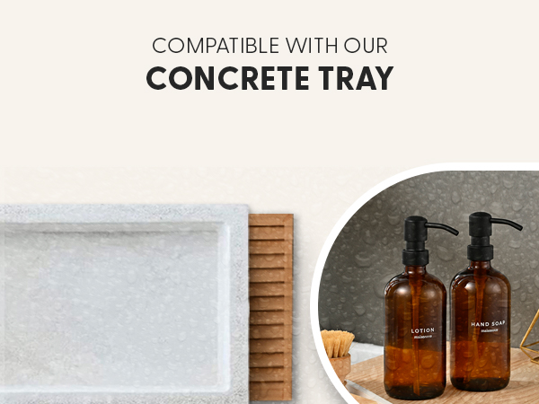 Text reads 'COMPATIBLE WITH OUR CONCRETE TRAY'. Image shows white concrete rectangular tray, wooden bamboo mat, and two dark amber glass pump bottles on orange/peach surface.