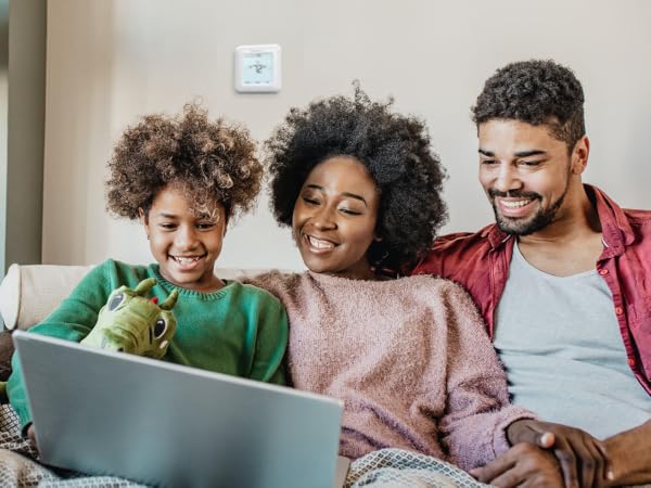 Family sitting on couch together using laptop computer, thermostat installed on background wall