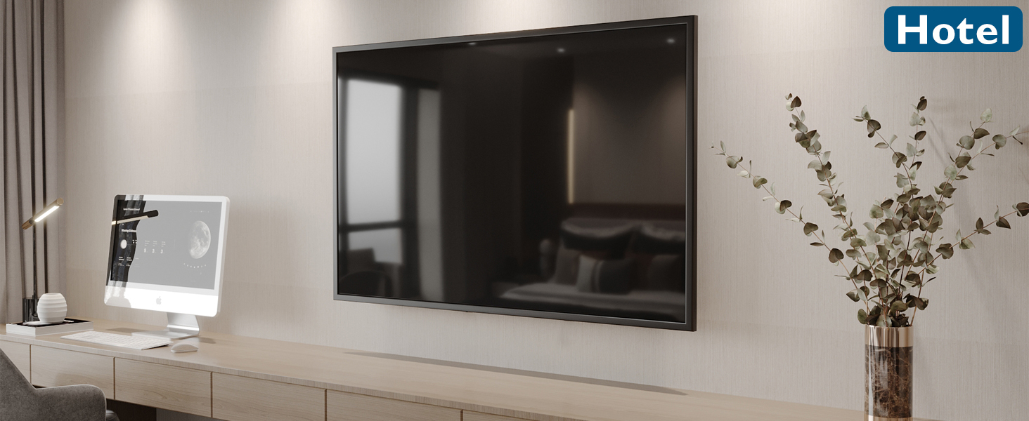 Modern hotel room with wall-mounted flat-screen TV, white computer on desk, and decorative plant. Clean, minimalist design with neutral tones.