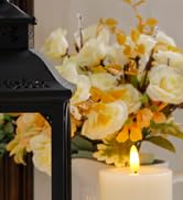 Decorative arrangement with white and yellow flowers in a vase next to a black lantern. A soft, warm glow from candles or lights illuminates the scene.