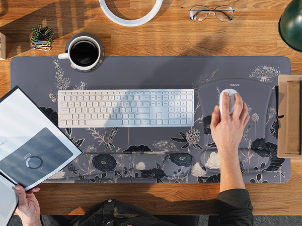 Overhead desk workspace showing keyboard, mouse, coffee cup, smartphone, and notebook on dark desk mat with geometric pattern.