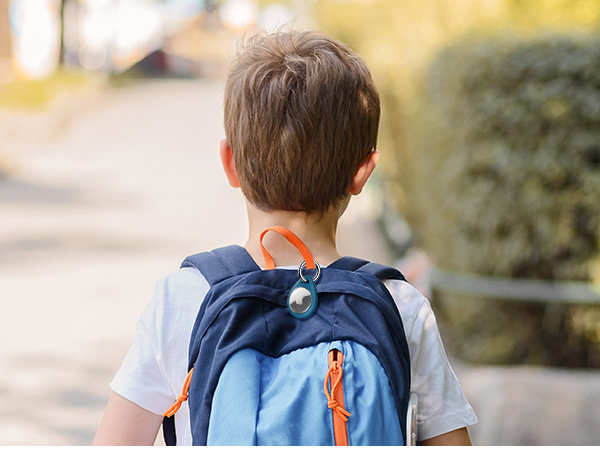 Child wearing a blue backpack with orange straps and a small plush toy attached, viewed from behind on a sunny day.