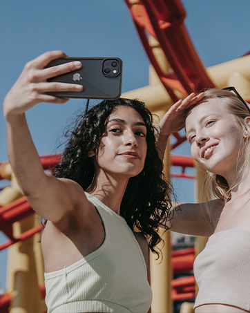 Black smartphone with dual cameras used to take a selfie outdoors. Red roller coaster structure visible in background.