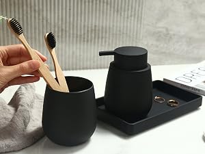 Matte black bathroom set featuring soap dispenser, toothbrush holder with wooden toothbrushes, and a tray holding jewelry. Hand placing toothbrush in holder.