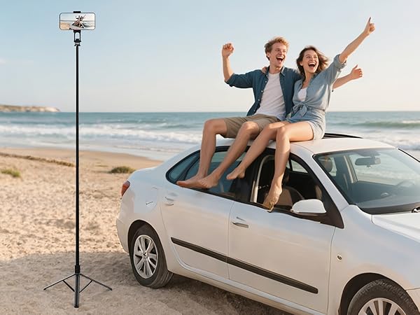 Extendable smartphone tripod stand on beach. Black metal pole with adjustable height, phone mount at top. White car and ocean visible in background.