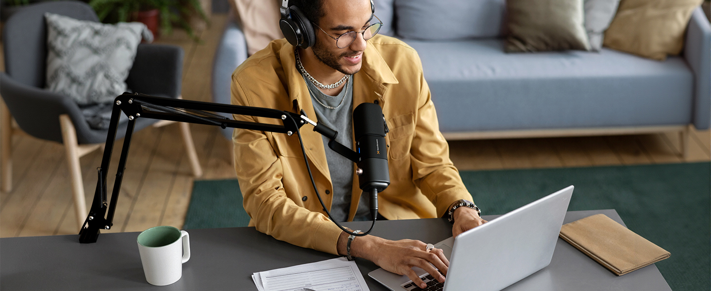 Person wearing tan jacket using laptop at desk with adjustable microphone arm and coffee mug, suggesting podcast or remote work setup.