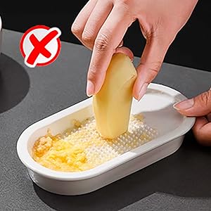 Hand grating potato on a small white grater with container. Red 'X' icon in corner suggests incorrect technique or safety warning for kitchen tool use.