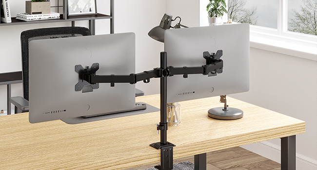 Dual monitor arm mount holding two gray computer monitors on a wooden desk. Adjustable arms for flexible positioning in an office setting.