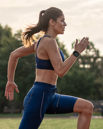 Woman in blue sports bra and leggings running outdoors, arms bent at elbows, ponytail swinging, against blurred green background.