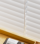 Close-up of white horizontal window blinds, partially opened to let light through. Mounted on a wooden window frame or sill.