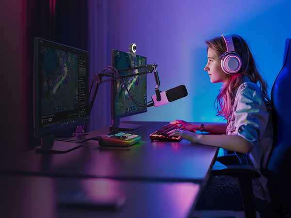 Young person wearing white and pink headphones sits at desk with gaming setup, viewing monitor in purple and blue neon-lit room environment with gaming peripherals visible.