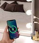 Illuminated vanity mirror with smartphone displaying charging icon, suggesting wireless charging capability integrated into the mirror's base.