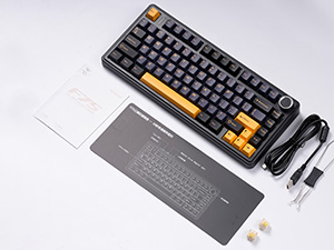 Compact mechanical keyboard with black and orange keycaps, shown with detached USB cable and additional accessories on light gray surface.