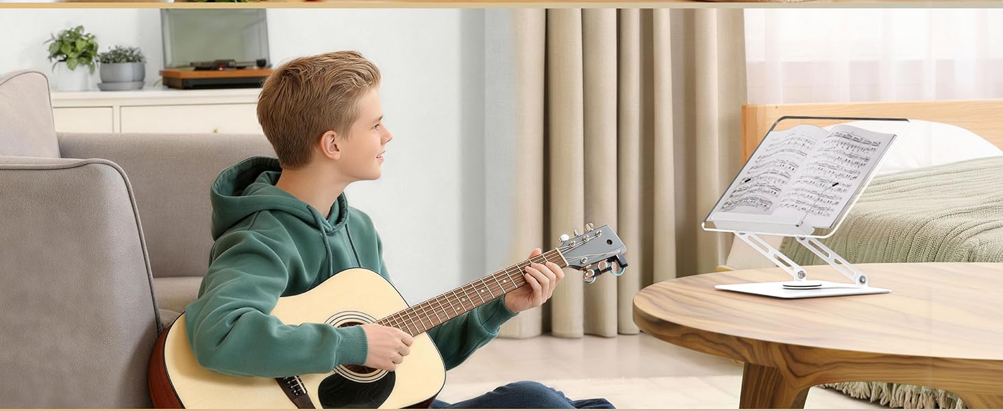Young person in green hoodie playing acoustic guitar while seated at desk with laptop stand and tablet holder setup.