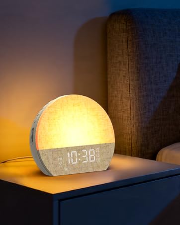 Digital alarm clock with glowing yellow/white display showing "19:38" time. Spherical white matte finish clock with warm LED lighting on dark blue nightstand with wooden post.