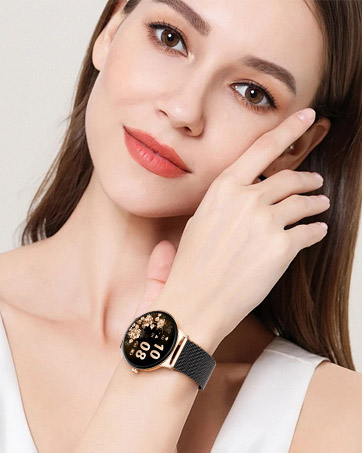 Round smartwatch with black and gold face displaying digital time. Black mesh band. Worn on wrist of person touching face.