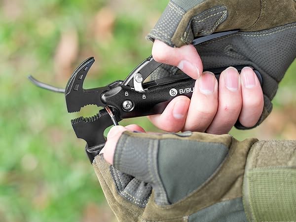 Close-up of tactical gloved hand holding compact black multi-tool or grappling hook device with curved metal prongs.