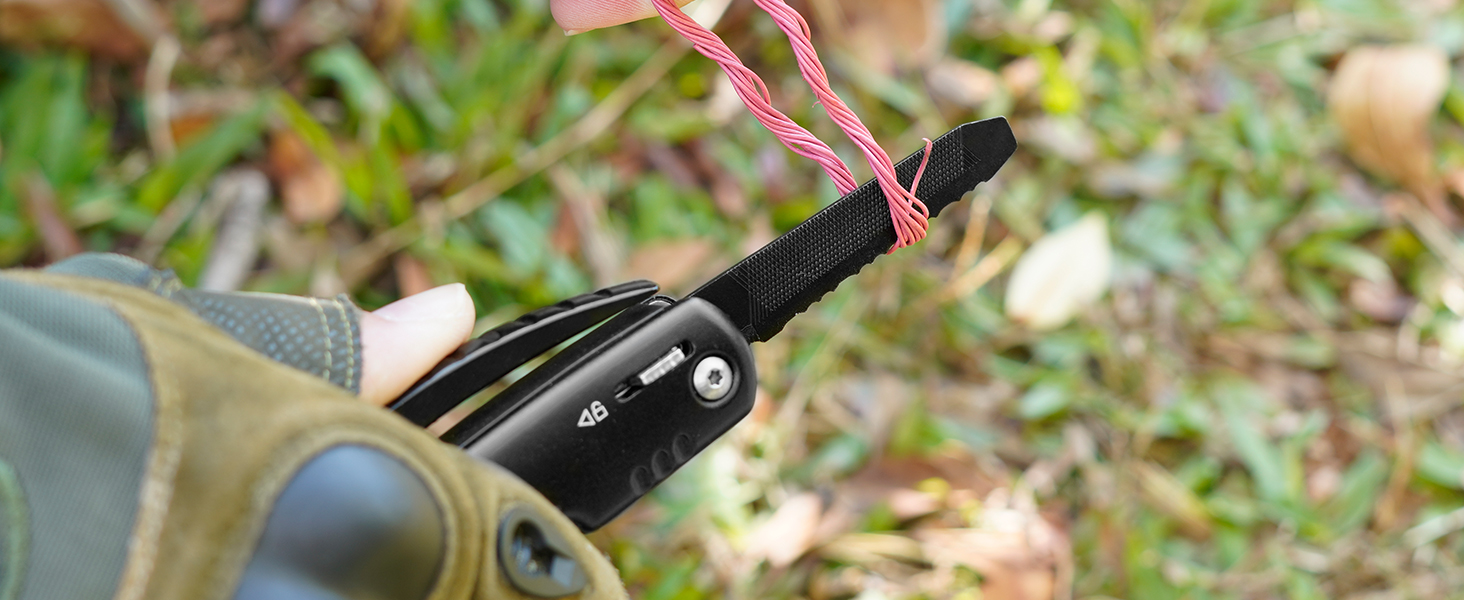 Close-up of a gloved hand holding a black folding knife with a pink cord attached. Background shows grass and leaves.