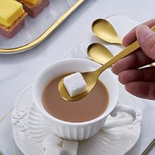Gold spoon dipping sugar cube into coffee cup. White porcelain cup and saucer on yellow surface. Partial view of additional items.