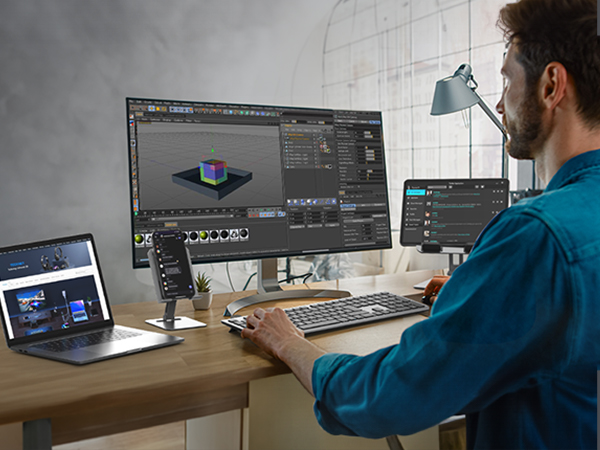 Workstation with multiple monitors displaying 3D modeling software. A person in a blue shirt is working at the desk with a laptop and desktop computer setup.
